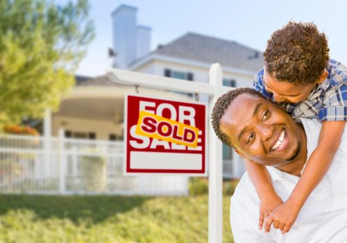 Happy African American Father and Mixed Race Son In Front of Sold Home For Sale Real Estate Sign and New House.